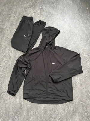 Svart Nike miler tracksuit storlek XL - Säljer ett svart nike miler set från Nike i storlek XL. Setet består av en jacka med huva och  samt matchande byxor, båda med vita Nike-loggor. Materialet är lätt och passar perfekt för löpning eller gym. Snygg och sportig look!