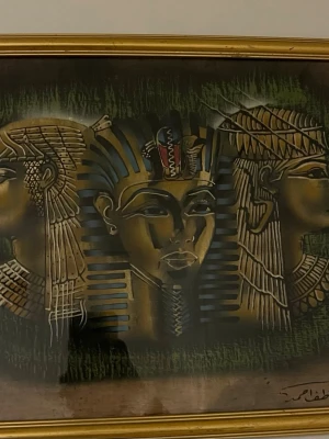 Tavla med egyptiskt motiv i guldram - Unik tavla med motiv av tre egyptiska figurer, inklusive en farao i mitten. Färgerna går i guld, svart, blått och grönt med detaljerade linjer och mönster. Tavlan är inramad i en tunn guldfärgad ram och har en signatur i hörnet.