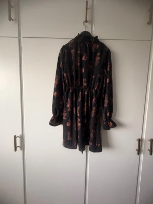 Svart blommig kortklänning med volang - Kort svart klänning från Gina Tricot med bruna blommor, hög krage och långa ärmar med volang vid ärmslut. Klänningen har resår i midjan för en snygg passform och är perfekt för dig som gillar en feminin och trendig stil.