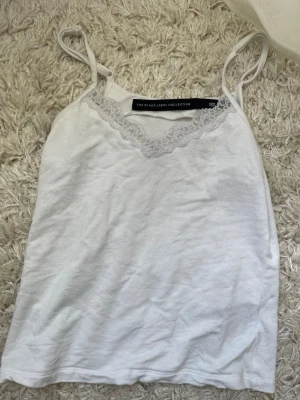 Vit spetsdetaljerad linnetopp  - Säljer en vit linnetopp från The Black Label Collection i storlek S. Toppen har tunna axelband och v-ringning med fina spetsdetaljer framtill. Perfekt att styla till jeans eller kjol för en fräsch look.