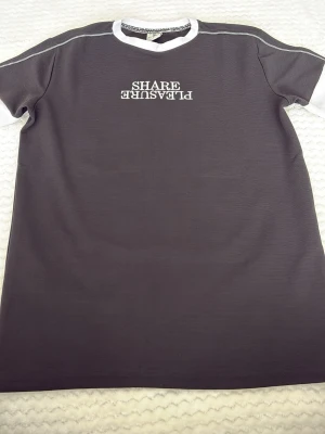 Svart t-shirt från E.Point - Cool svart t-shirt från E.Point med vit text 'SHARE PLEASURE' broderad på bröstet, där 'PLEASURE' är upp och ner. T-shirten har vita kontrastdetaljer vid krage och ärmslut samt synliga sömmar på axlarna. Perfekt för en avslappnad streetwear-look.