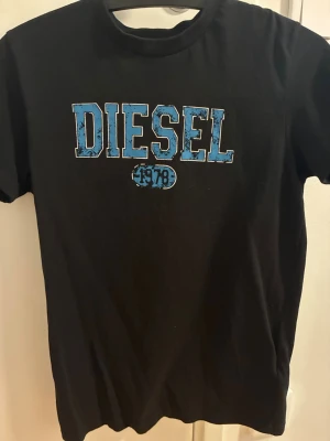 Svart t-shirt från Diesel med tryck - Snygg svart t-shirt från Diesel med stort blått och vitt DIESEL-tryck och årtalet 1978 på bröstet. Klassisk rund hals och korta ärmar. Perfekt för dig som gillar streetwear och vill ha en enkel men cool look.