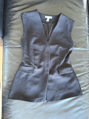 Svart väst från H&M med v-ringning - Stilren svart väst från H&M i storlek XS. Västen har en snygg v-ringning, figurnära passform och dekorativa fickor framtill. Perfekt att styla över en skjorta eller t-shirt för en trendig look.