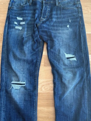 Jack and jones jeans  - Säljer ett par mörkblå jeans med slitningar och hål på både lår och knän. Jeansen har klassisk femficksmodell, raka ben och tydliga slitdetaljer för en trendig look. Perfekta för dig som gillar en avslappnad och edgy stil.