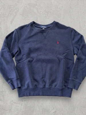 Mörkblå sweatshirt från Polo Ralph Lauren - Mörkblå sweatshirt med rund hals och långa ärmar från Polo Ralph Lauren. Klassisk design med ribbade muddar och diskret röd logobrodyr på bröstet. Perfekt för en avslappnad och stilren look. Det är en barn storlek L så passar kanske XS och S. Ni får mer än gärna skriva om mer info eller lägga bud😊