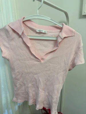 Ljusrosa ribbad polotopp från Stradivarius  - Söt ljusrosa ribbad topp från stradivarius med krage och v-ringning. Kortärmad och croppad modell som ger en trendig look. Perfekt att matcha med jeans eller kjol för en avslappnad stil.