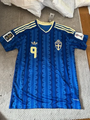 Blå Sverige retro matchtröja Adidas - Retroinspirerad blå matchtröja från Adidas med Sveriges landslagsemblem och nummer 9 på bröstet. Tröjan har gula detaljer, klassiska Adidas-ränder på axlarna och ett unikt mönster i tyget. Perfekt för dig som älskar fotboll och vill sticka ut på läktaren.