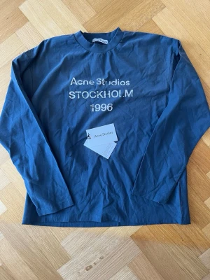 Blå långärmad t-shirt Acne Studios - Snygg blå långärmad t-shirt från Acne Studios med tryck 'Acne Studios STOCKHOLM 1996' på bröstet. Klassisk rund hals och relaxed fit. Tillverkad i bomull och true hemp, perfekt för en clean och stilren look.