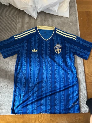 Blå Sverige fotbollströja Adidas - Säljer en blå fotbollströja från Adidas med Sveriges landslagsemblem på bröstet. Tröjan har gula detaljer, klassiska Adidas-ränder på axlarna och ett unikt mönster över hela tyget. Perfekt för dig som vill visa ditt stöd för Sverige på matchen!