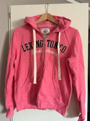 Rosa hoodie från Lexington - Snygg rosa hoodie från Lexington med dragkedja, vit text på bröstet och stora fickor framtill. Mjukt material, huva med vita snören och ribbade muddar. Perfekt för en avslappnad och trendig stil.