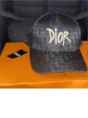 Svart Dior keps med logotyp - Snygg svart keps från Dior med tydligt mönster av Dior-loggor över hela kepsen och stor broderad logotyp framtill i beige. Klassisk böjd skärm och justerbar baksida. Perfekt accessoar för att lyfta din streetwear-look.
