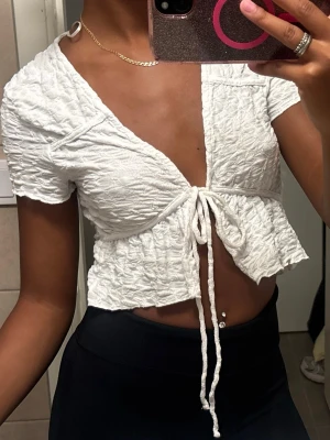 Vit croppad topp med knytning - Trendig vit croppad topp med korta ärmar och djup v-ringning. Toppen har ett snyggt rynkat tyg och knytning framtill som ger en cool och avslappnad look. Perfekt till höga byxor eller kjol för en modern stil.