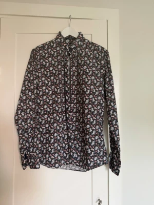 Blommig långärmad skjorta - Snygg långärmad skjorta med blommigt mönster i vitt och rosa på svart botten. Skjortan har klassisk krage och knappar framtill. Perfekt för dig som vill sticka ut lite extra med en färgglad och trendig look.