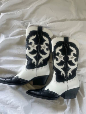 Svarta och vita cowboy boots med stjärnor - Säljer ett par riktigt snygga cowboy boots i svart och vitt med coola stjärndetaljer och kontrastsömmar. Bootsens skaft har ett unikt mönster och bootsen har spetsig tå samt bred klack. Perfekta för dig som vill sticka ut med din stil.