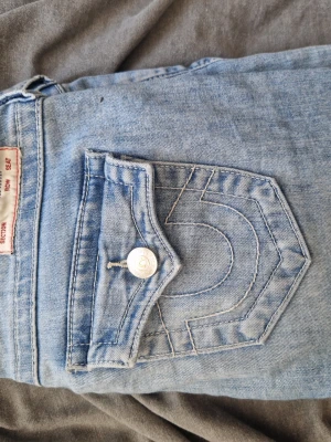Ljusblå True Religion jeans - Säljer ett par ljusblå jeans från True Religion med klassisk kontrastsöm och ikonisk fickdesign med lock och knapp bak. Jeansen har låg midja, raka ben och är tillverkade i bomull. Perfekta för en avslappnad och trendig look. Säljer eftersom dem inte passar.
