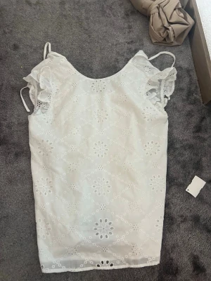 Vit broderad blus med volangärmar - Söt vit blus med broderade blommor och små hål, volangdetaljer vid ärmarna och smala axelband. Perfekt för sommarens varma dagar och ger en romantisk känsla till din outfit.