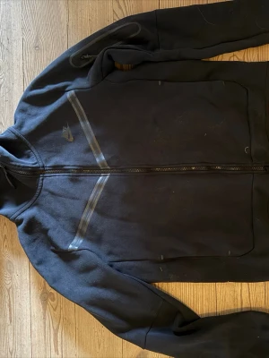 Svart Nike OG Tech Fleece  - Säljer en svart Tech Fleece från Nike med hel dragkedja framtill och två snedställda fickor. Hoodien har en diskret Nike-logga på bröstet och tunna gråa detaljer över bröstet. Tillverkad i mjukt bomullsmaterial och har huva.