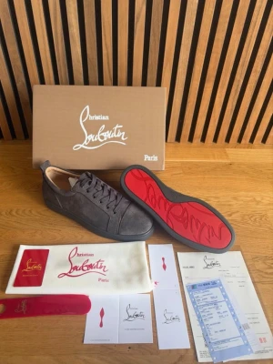 Christian Louboutin gråa sneakers - Snygga gråa sneakers från Christian Louboutin med klassisk röd sula och lågt skaft. Skorna är tillverkade i mocka och har svarta skosnören. Perfekta för dig som vill ha en exklusiv och stilren look med den ikoniska röda sulan. Storlek 43! Vid frågor eller funderingar är de bara att höra av sig! 
