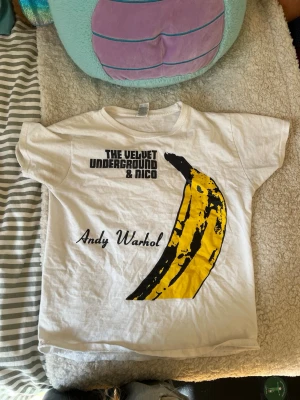 Vit The Velvet Underground t-shirt - Säljer en vit t-shirt med ikoniskt Andy Warhol-banantryck och texten 'The Velvet Underground & Nico'. T-shirten har korta ärmar och är i mjuk bomull, perfekt för dig som gillar klassiska bandmotiv och konstnärlig stil. (Den har pyttesmå deofläckar som knappt syns)