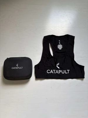 Catapult GPS-väst - Säljer en svart GPS-väst från Catapult i storlek M, perfekt för att mäta prestation vid träning. Västen är i syntetmaterial och kommer med en hård förvaringsväska samt tillhörande sensor. Allt är svart och har tydlig Catapult-logga.