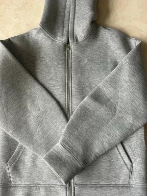 Grå zip hoodie med fickor - En stilren grå hoodie med dragkedja framtill och två stora fickor. Hoodien har huva och ribbade muddar vid ärmsluten. Tillverkad i mjukt sweatshirtmaterial som känns skönt mot huden. Perfekt för en avslappnad och casual look. Goodwin är lite större i storleken.