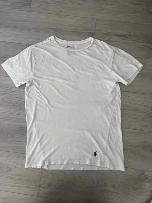   Polo Ralph Lauren T-shirt - Klassisk vit t-shirt från Polo Ralph Lauren med den ikoniska mörkblå broderade loggan vid nederkanten. T-shirten har rund halsringning och korta ärmar, perfekt för en clean och stilren look.