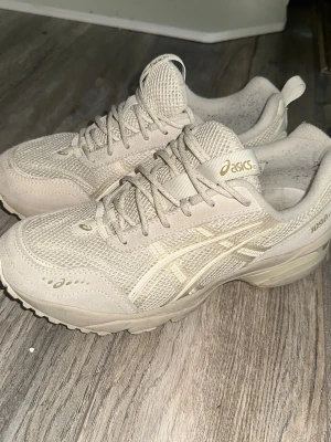 Beige Asics sneakers mesh och mocka - Snygga beige sneakers från Asics med ovandel i mesh och mocka. Skorna har klassisk Asics-logga på sidan och tungan, samt grov sula för extra komfort. Perfekta för dig som gillar stilrena och neutrala färger.