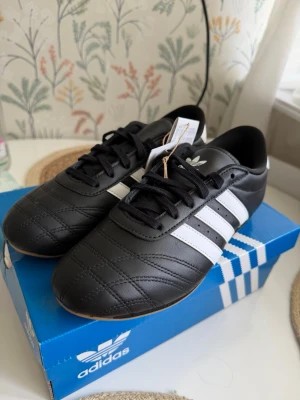 Adidas TEAKWONDO size 38 - Klassiska Adidas sneakers i svart läder med vita detaljer och de ikoniska tre ränderna på sidan. Låg modell med svart snörning och vit häl med Adidas-logga. Snygg retrostil som passar till många outfits. Yttersulan är i gummi för bra grepp.