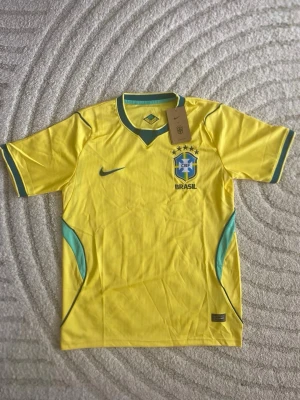 Gul Brasil Nike fotbollströja - Snygg gul fotbollströja från Nike med Brasiliens landslagslogga och fem stjärnor på bröstet. Tröjan har gröna detaljer vid krage och ärmslut samt blå inslag vid sidorna. Tillverkad i lätt och ventilerande material, perfekt för träning eller match.