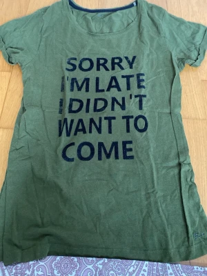 Grön t-shirt med texttryck - Snygg grön t-shirt med rund halsringning och korta ärmar. Framsidan har ett stort svart texttryck: 'SORRY I'M LATE I DIDN'T WANT TO COME'. Tillverkad i mjukt bomullstyg, perfekt för en avslappnad stil.