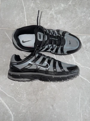 Nike P-6000 svart/grå sneakers - Nike P-6000 sneakers i svart och grå mesh med svarta syntetiska överdrag och vit Swoosh på sidan. Skorna har en chunky sula och sportig design med snörning och detaljerade paneler. Perfekta för dig som gillar retrovibbar och streetwear.