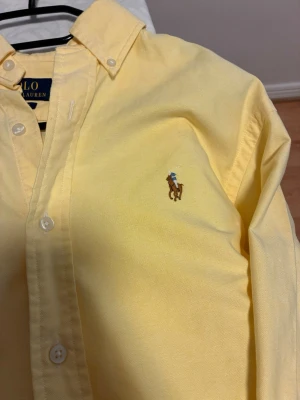 Gul skjorta från Polo Ralph Lauren - Klassisk gul skjorta från Polo Ralph Lauren med broderad logga på bröstet. Skjortan har knappar framtill och button-down krage. Tillverkad i bomull och har en normal passform. Perfekt för en clean och stilren look.