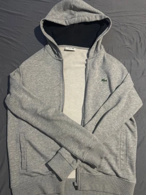 Grå zip hoodie från Lacoste - Grå zip hoodie från Lacoste. Modellen är Lacoste sport så passformen är perfekt till den som bär M eller S. Materialet är riktigt mjukt och skönt måste jag säga. Perfekt till någon mellan 175-185 och 65-80kg. Tröjan är använd ett fåtal gånger då den inte varit så viktig för mig. Topp skick verkligen. Inga tecken på användning utom att jag tagit av snörena.