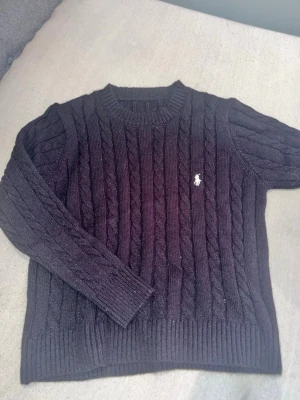 Svart stickad tröja Polo Ralph Lauren - Womans stickad svart tröja från Polo Ralph Lauren med klassisk rund halsringning och vit broderad logga på bröstet. Tröjan har långa ärmar och ribbade muddar vid ärmslut och nederkant.