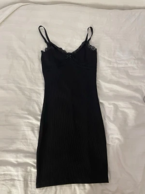 Svart ribbat nattlinne med spets - Snyggt svart nattlinne/bodycon dress med tunna axelband och ribbad struktur. Dekorerat med spetsdetaljer vid urringningen och en liten rosett framtill. Mjukt och stretchigt material som känns bekvämt mot huden. Perfekt för mysiga kvällar hemma.