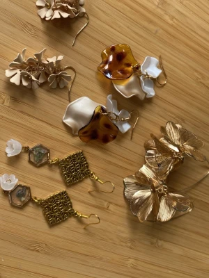 Mixade statementörhängen med blommor - Säljer ett set med fyra par unika statementörhängen. Blandade material i guld, beige, vitt och sköldpaddsmönstrat brunt. Flera örhängen har blommotiv i metall och plast, samt detaljer som geometriska former och små pärlliknande inslag. Perfekt för dig som gillar att sticka ut.