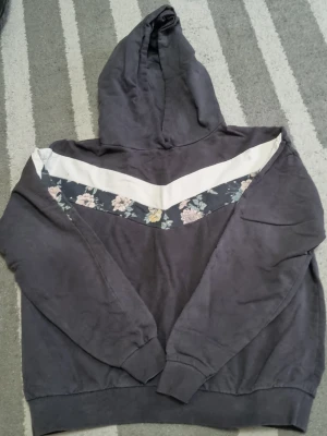 Mörkgrå hoodie med blommigt band - Snygg mörkgrå hoodie med huva och ett vitt och blommigt band över bröstet. Hoodien har långa ärmar, ribbade muddar och en avslappnad passform. Perfekt för en chill och trendig look.