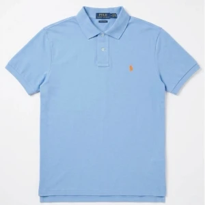 Blå pikétröja från Polo Ralph Lauren - Klassisk blå pikétröja från Polo Ralph Lauren med den ikoniska broderade loggan i orange på bröstet. Tröjan har krage, korta ärmar och knappar vid halsen. Perfekt för en avslappnad men ändå stilren look.