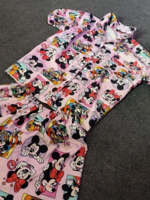 Rosa Disney pyjamas med Musse & Mimmi - Säljer en söt tvådelad pyjamas i rosa med färgglada tryck av Musse och Mimmi Pigg. Skjortan har korta ärmar, krage och knappar framtill. Byxorna har resår i midjan för extra komfort. Perfekt för dig som älskar Disney och vill ha något bekvämt att sova i.