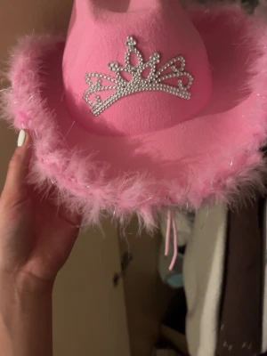 Rosa cowboyhatt med tiara-detalj - Säljer en ikonisk rosa cowboyhatt med fluffig fjäderkant och en glittrig tiara i strass framtill. Hatten är gjord i mjukt filtmaterial och har en riktigt statement look som passar perfekt för temafester eller festivaler.