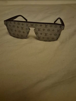 Lv solglasögon i monogram glas - Snygga solglasögon med spegelglas i silver och ikoniskt monogrammönster över hela linsen. Bågarna är svarta och modellen har en futuristisk, kantig form som sticker ut. Perfekta för dig som vill ha en exklusiv och trendig look.