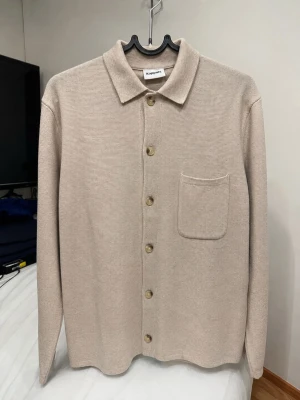 Beige stickad tröja från Kappahl - Säljer en stilren beige stickad kofta från Kappahl med klassisk krage, knappstängning och en bröstficka. Koftan har lång ärm och är tillverkad i ett mjukt, tunt material som passar perfekt för lager på lager.