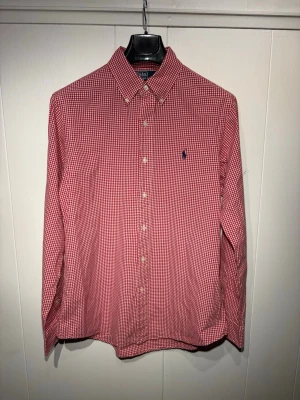 Röd vit rutig skjorta från Polo Ralph Lauren - Klassisk röd och vit rutig skjorta från Polo Ralph Lauren med button-down krage och broderad logga på bröstet. Skjortan är tillverkad i mjuk bomull. Perfekt nu under sommaren med de röda och vita som bjuder på en ljus skön look. Passar till både kavaj men även till att bara hänga en tröja över axlarna. Skjortan är i fläckfritt skick och har inte några defekter.
