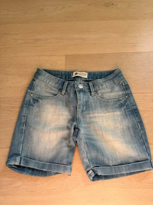 Blå jeansshorts från ICHI - Säljer ett par klassiska blå jeansshorts från ICHI med fem fickor och uppvikta benslut. Shortsen har normal passform och stängs med knapp och dragkedja. Perfekta till sommaren och i bomull för en skön känsla.