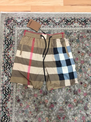 Rutig shorts från Burberry - Säljer ett par klassiska rutig shorts från Burberry med beige, svart, vit, blå och röd färg. Shortsen har snörning i midjan och elastisk linning för skön passform. Perfekta för sommaren och har den ikoniska Burberry-looken. Oanvända burberry shorts i storlek M