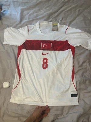 Turkiet matchtröja Nike Arda Güler - Snygg vit och röd matchtröja från Nike med Turkiets flagga på bröstet och nummer 8. På ryggen står det 'Arda Güler' och siffran 8 i rött. Tröjan har korta ärmar, V-ringning och är tillverkad i ett lätt, ventilerande material perfekt för fotboll. Är storlek S men sitter mer som M