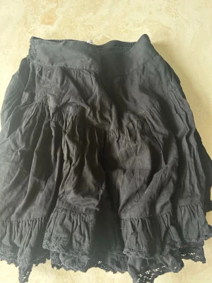 Svart volangkjol med spetsdetaljer - Kort svart kjol med flera volanger och fina spetsdetaljer längst ner. Kjolen är gjord i ett mjukt tyg. Den är lowwaist och har en väldigt snygg passform.