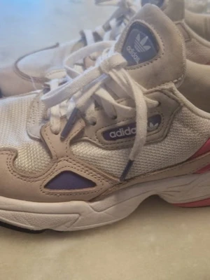 Adidas Falcon beige/lila sneakers - Snygga Adidas Falcon sneakers i beige mesh och mocka med lila och rosa detaljer. Klassisk chunky sula och retrovibbar. Skorna har snörning och Adidas-logga på plösen och sidan. Perfekta för dig som gillar streetstyle och sportig look.