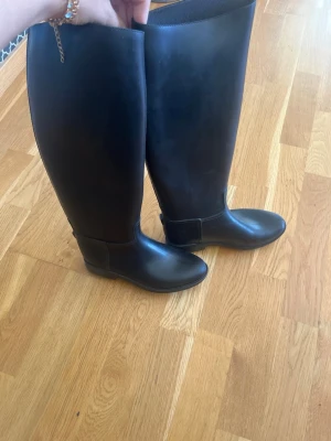 Svarta höga ridstövlar i gummi - Säljer ett par klassiska svarta ridstövlar i gummi med rund tå och slät sula. Stövlarna är knähöga och har en stilren, enkel design utan synliga detaljer eller dekorationer. Perfekta för regniga dagar eller till stallet.