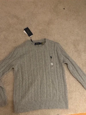  Ralph Lauren Sweatshirt - Kabelstickad grå tröja från Polo Ralph Lauren med rund halsringning och broderad logga på bröstet. Tröjan har långa ärmar och ribbade muddar vid ärmslut och nederkant. Perfekt för lager-på-lager och klassisk stil. Har aldrig använt endast packar upp och prövat.